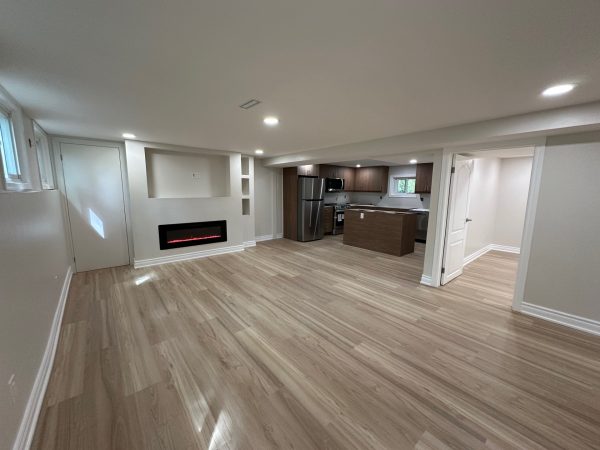 Open-concept finished basement with light wood flooring, recessed lighting, electric fireplace, and modern kitchenette.
